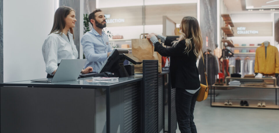 Clothing Store Checkout Cashier Counter: Beautiful Young Woman Buys Blouse from Friendly Retail Sales assistant, Receiving Bag with Purchases. Trendy Fashion Shop with of Designer Brands